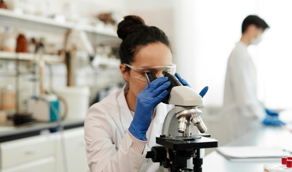 A laboratory scientist is examining a sample through a microscope while another researcher works in the background, representing medical research and diagnostic testing.