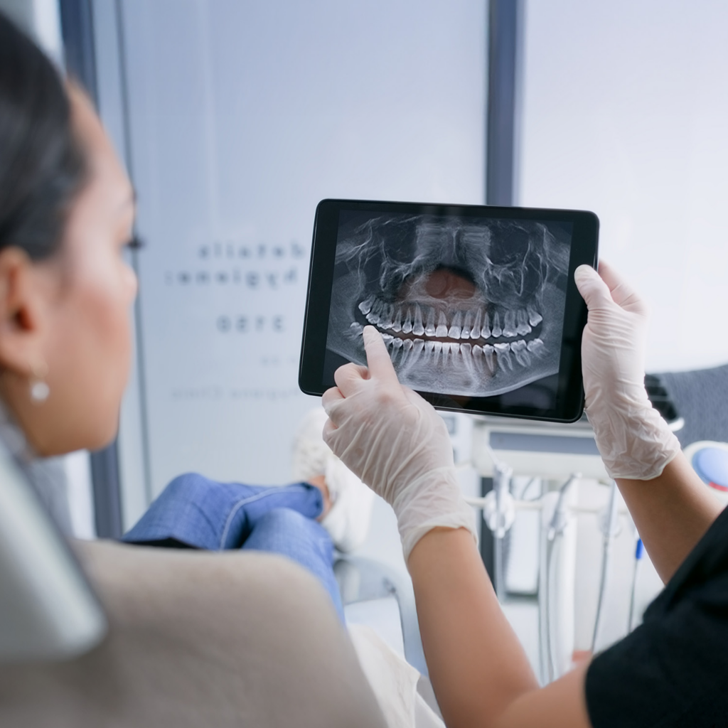 A dental professional is showing a patient a panoramic X-ray of their teeth on a tablet during a dental consultation.