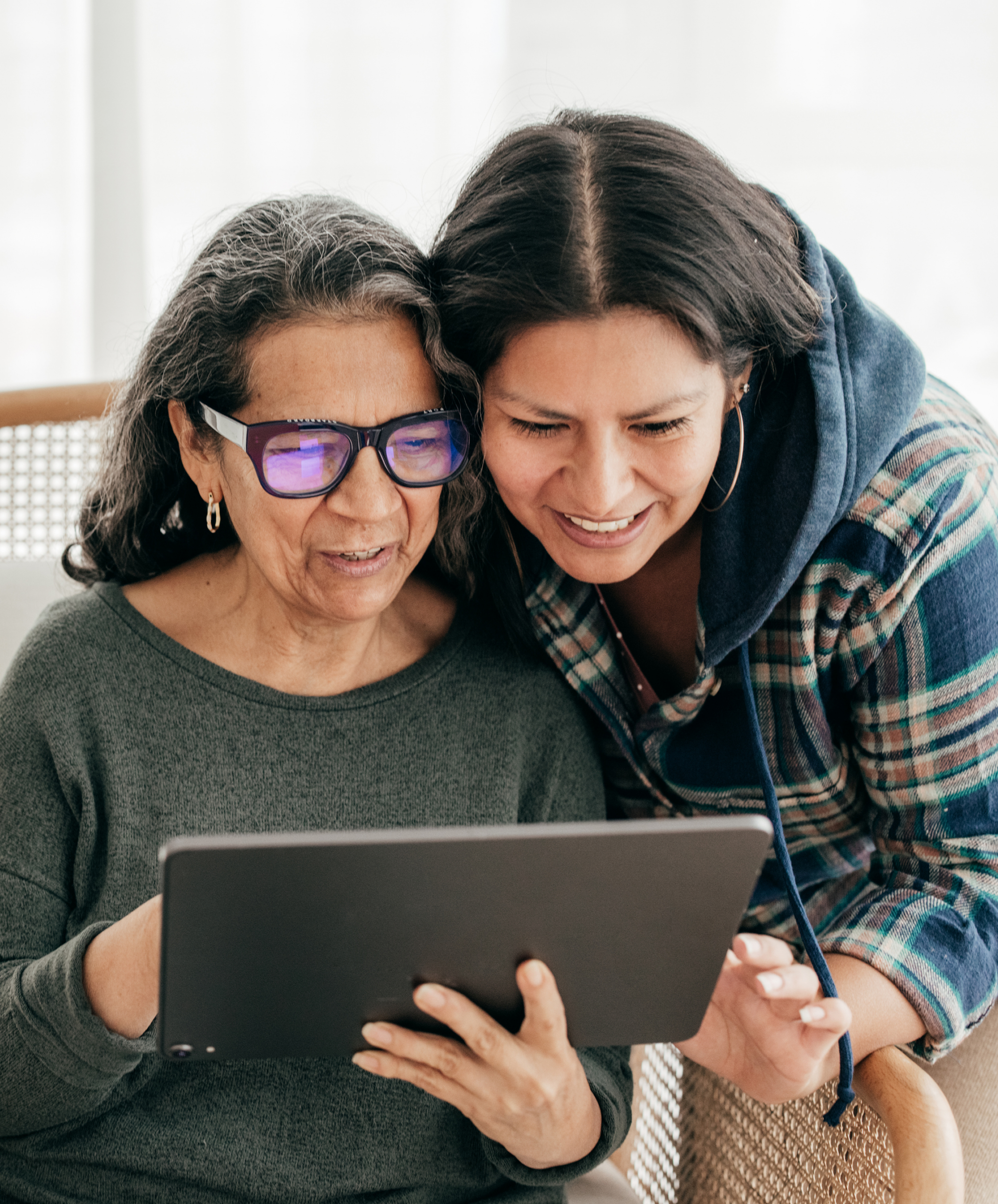 An older woman and a younger woman are smiling while looking together at a tablet screen, suggesting support, guidance, or access to digital healthcare information.